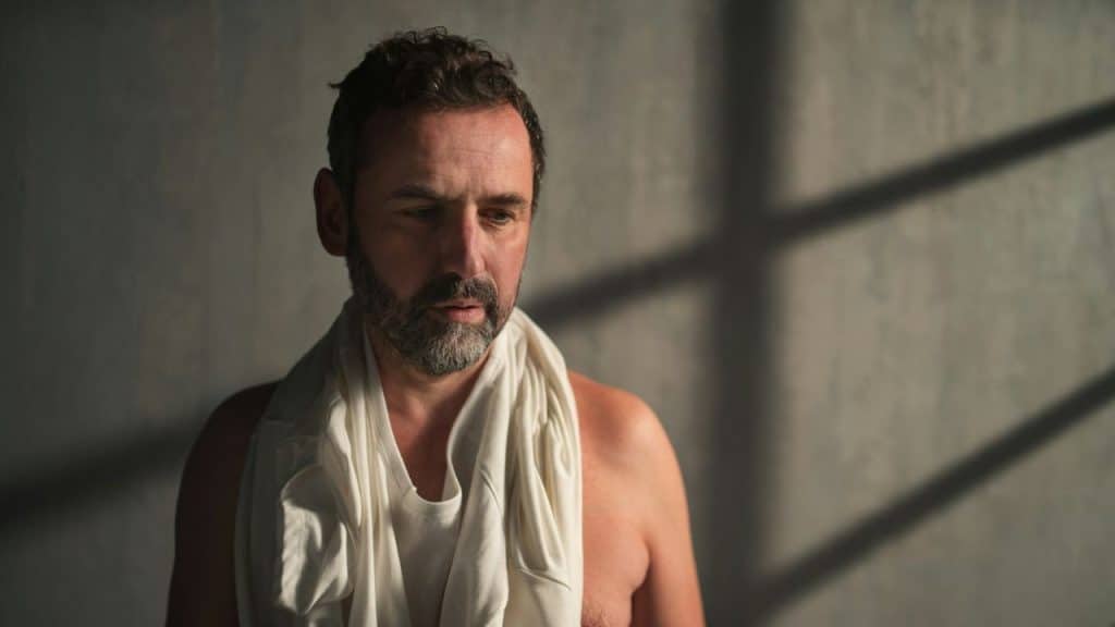 A man with a beard looks down pensively while holding a white shirt around his neck.