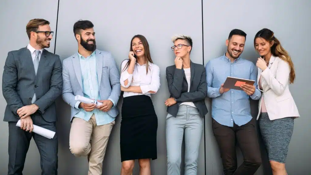 Six professionals in business attire stand against a grey wall, talking, laughing, and using devices.