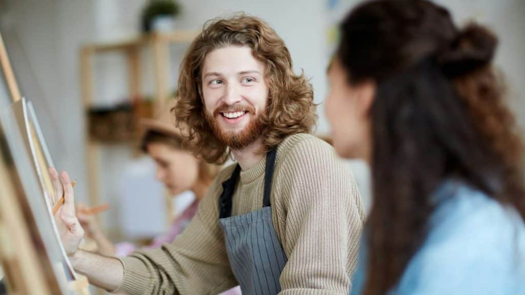 A man with long curly hair wears an apron and smiles while painting at an easel.