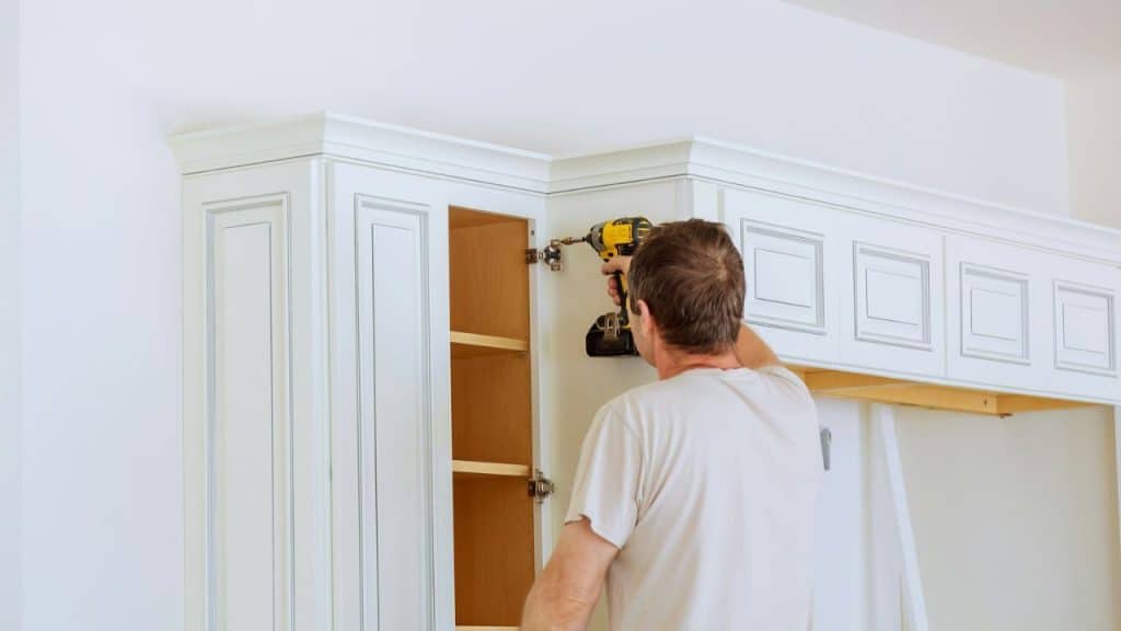 A man in a light-colored t-shirt uses a power drill to install white kitchen cabinets.