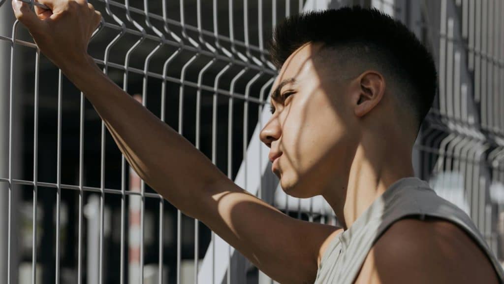 A person standing by a metal fence, looking outward in bright sunlight.