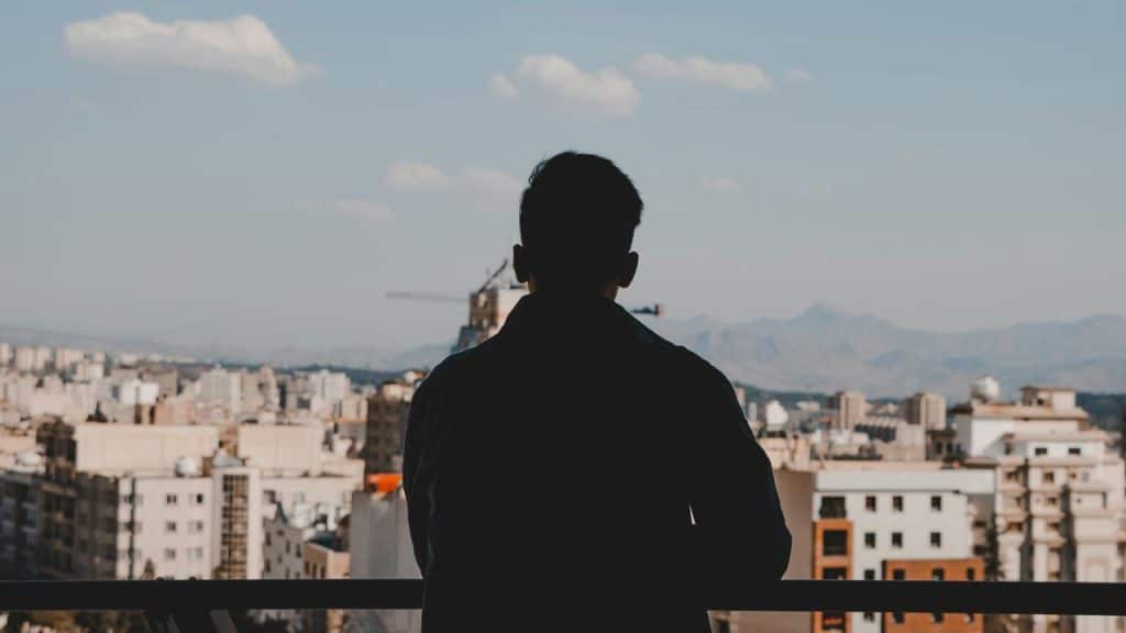 A man standing on a balcony looking out over a cityscape with mountains in the distance.