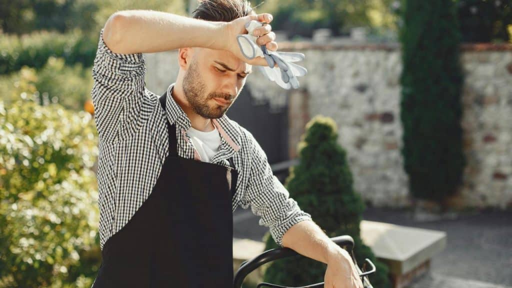 A man outdoors wearing an apron and wiping his forehead with work gloves.