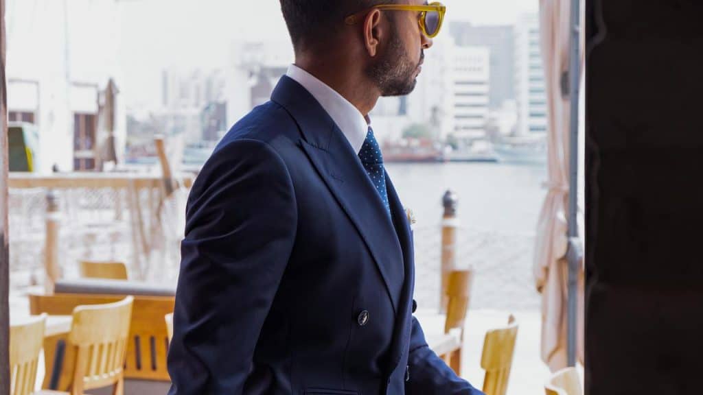 A man in a navy suit and yellow sunglasses looks outside near a waterfront.