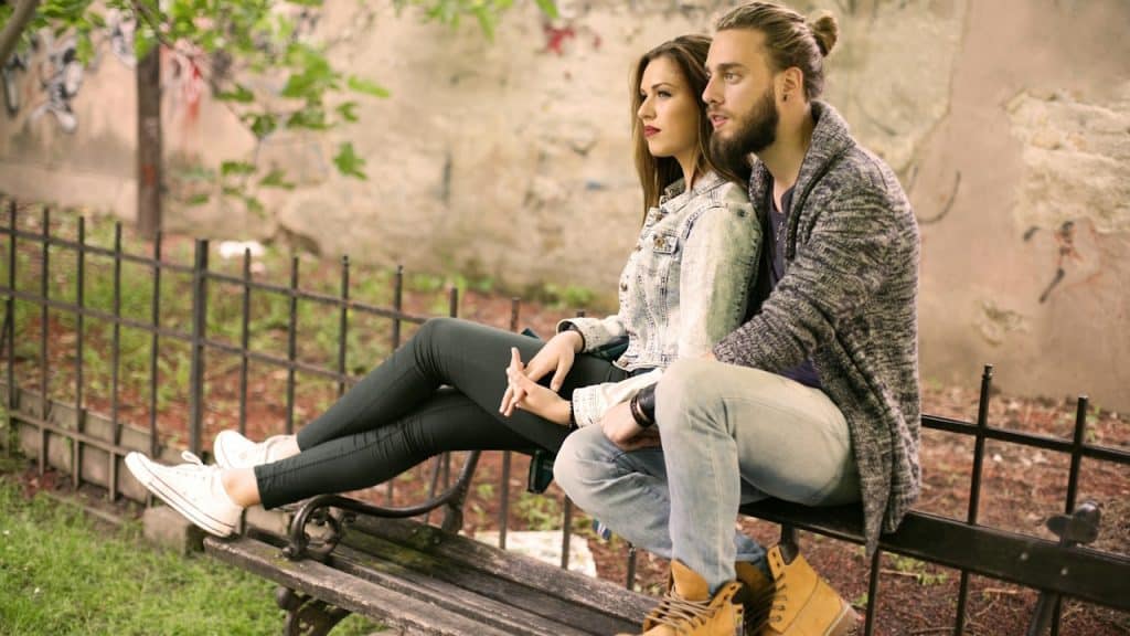 A couple sitting on a bench in front of a fence while the man holds the woman tightly as she leans on his chest.