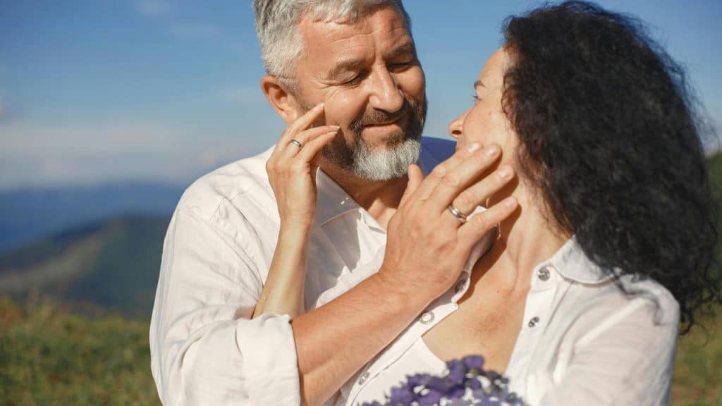 A couple smiling affectionately at each other on a sunny hillside.