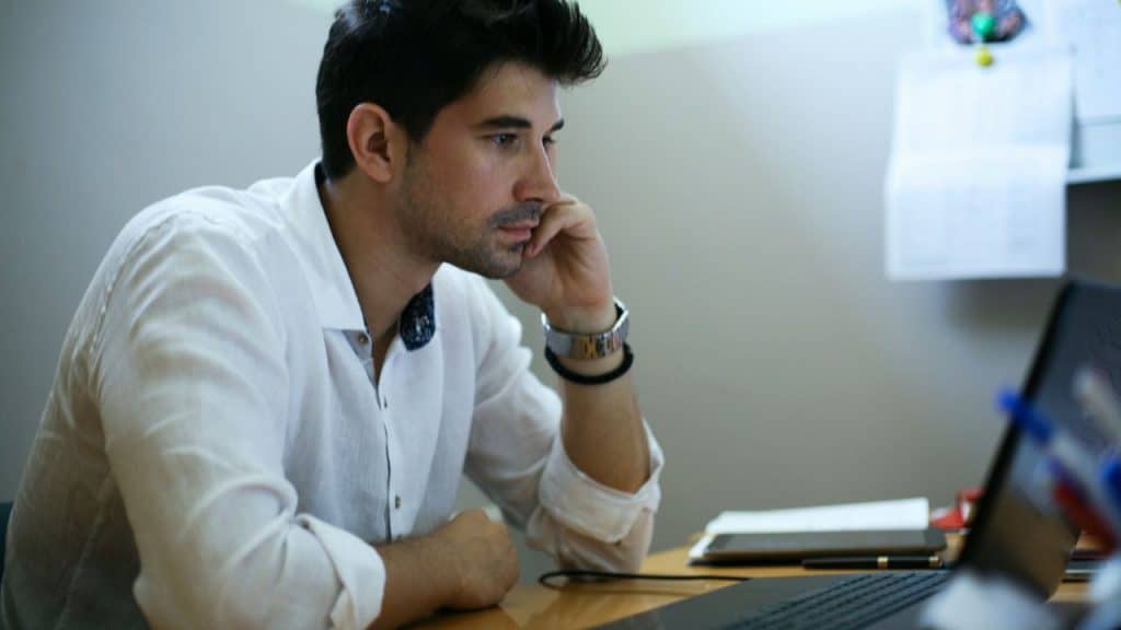 A man in a white shirt looks intently at a laptop screen with a serious expression.