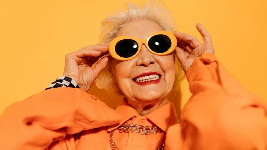 Senior woman with grey hair dressed in fashionable clothes smiling at the camera and holding hands at her sunglasses while posing at the studio with a yellow wall.