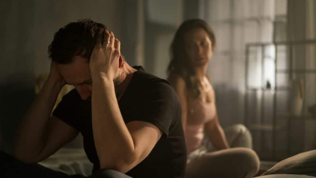 A man sits in the foreground with his head in his hands, looking distressed, while a woman sits blurred in the background on the bed.