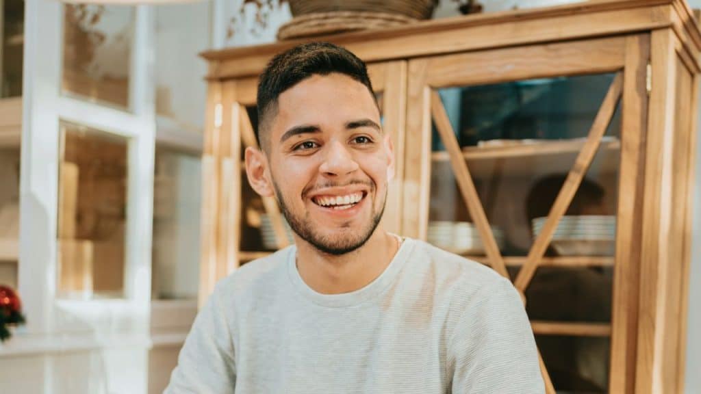 A smiling young man in a light grey shirt sits in front of a wooden cabinet.