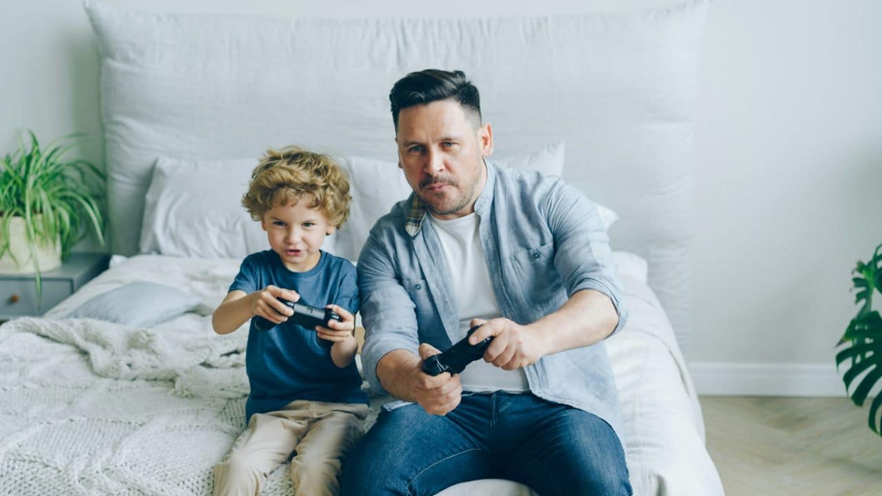 Father and son sitting on a bed, focused on playing a video game together.