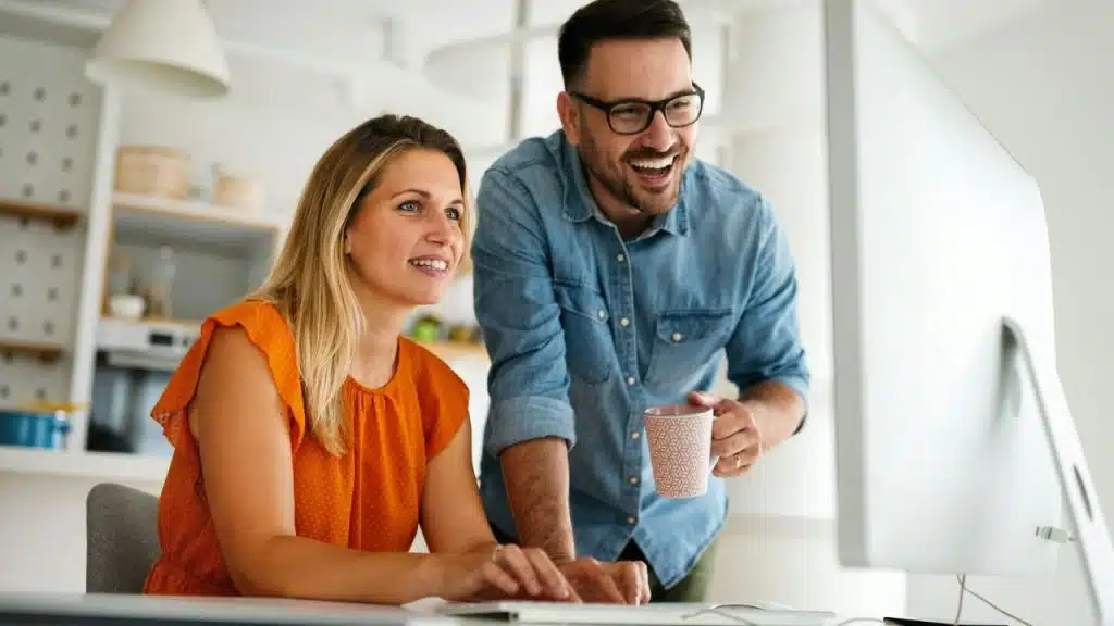 A smiling couple looks at a computer screen in a bright, modern office or kitchen setting.