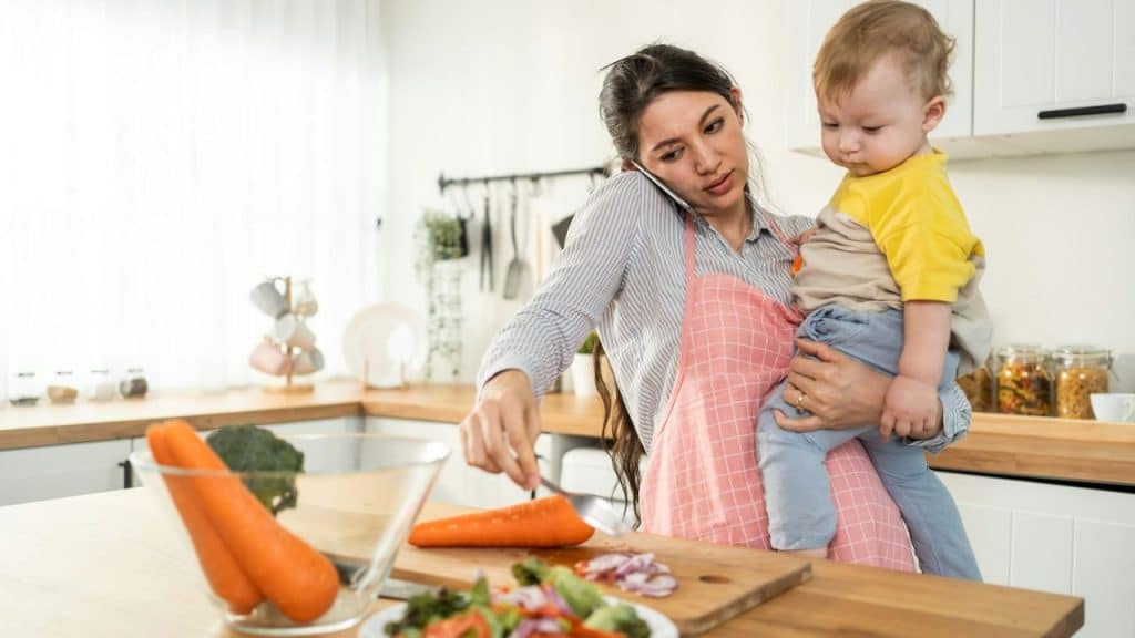 Woman in an apron holds a baby, talks on the phone, and chops a carrot in the kitchen.