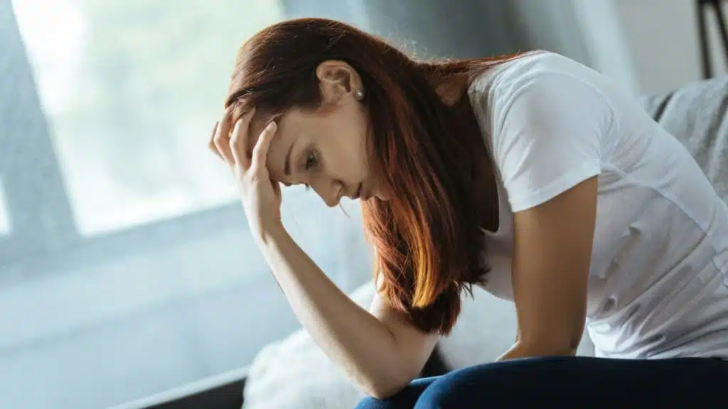 Woman with reddish-brown hair, wearing a white shirt, sitting with her head in her hand.