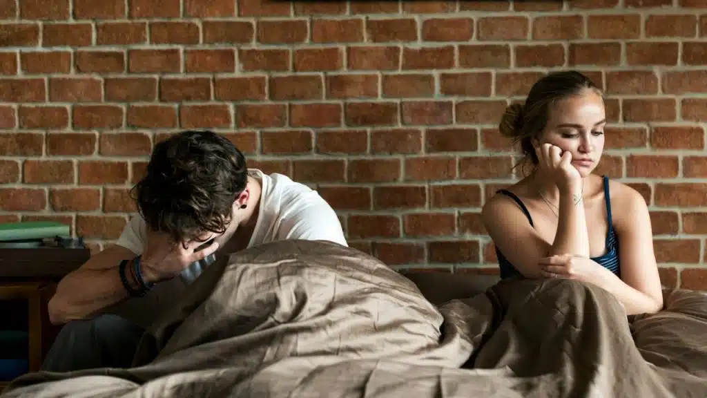 A man and woman sit apart on a bed, both looking upset against a brick wall.