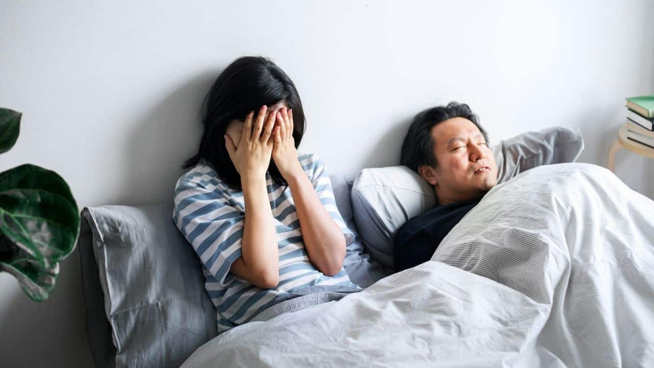 A woman sitting up in bed, covering her face with her hands, next to a sleeping man.