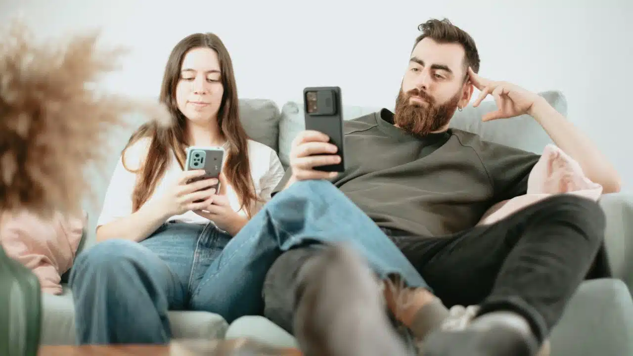 A man and a woman sit on a couch, both focused on their smartphones.