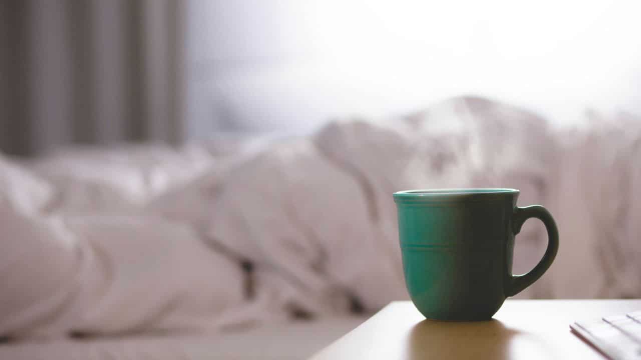 A steaming teal mug sits on a table with a blurred, white-blanketed bed in the background.
