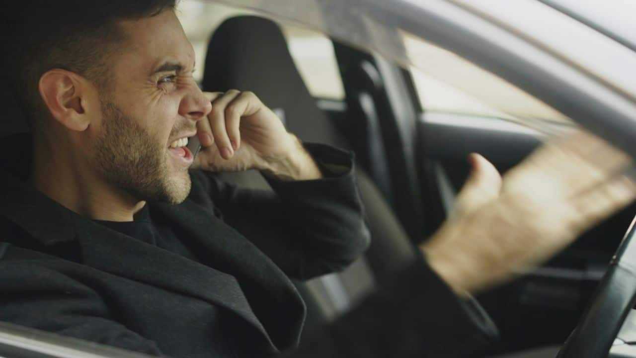 Angry man with short hair and stubble shouting while driving a car.