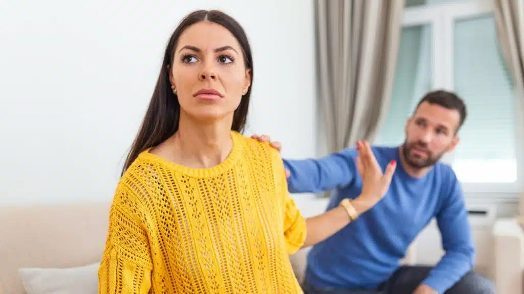 A woman in a yellow top looks away as a man reaches out to her shoulder.