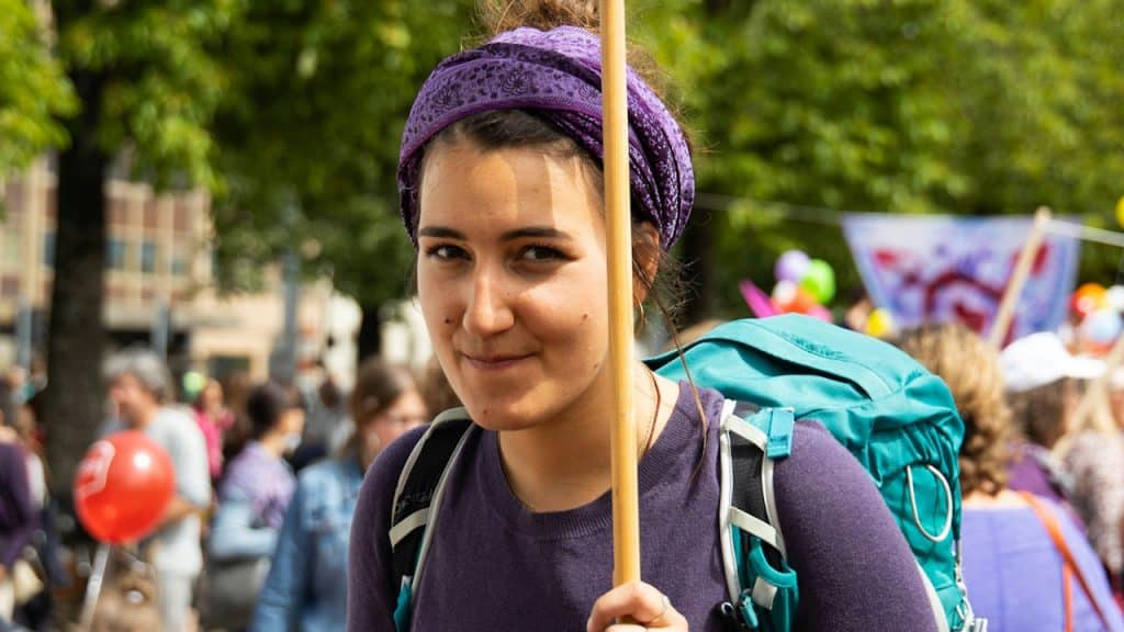 A woman wearing a purple dress and a scarf is holding a banner in a crowd.