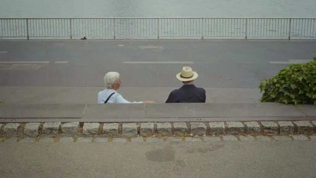 A couple sitting apart from each other on a bench by the street along the sea.