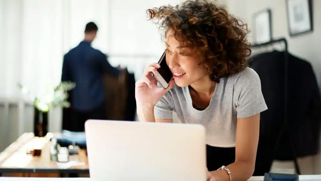 A woman talking on a phone while using a laptop as she sits in an office.