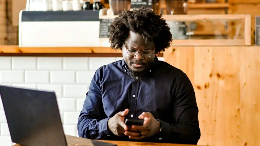 A man wearing glasses looking at his phone’s screen while his laptop lies open on a table in front of him.