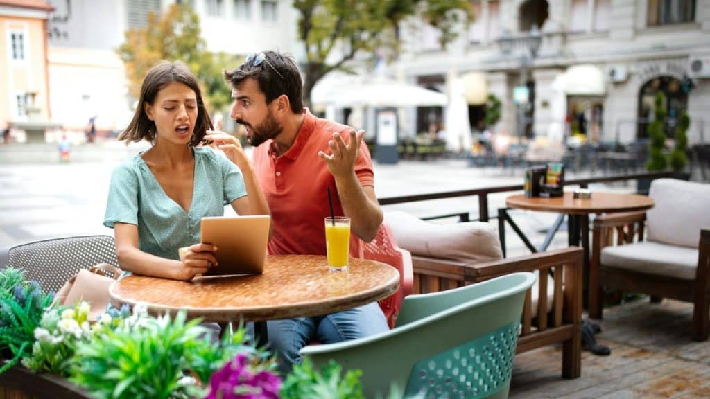 Man and woman arguing outdoors at a cafe table with a tablet and drink.