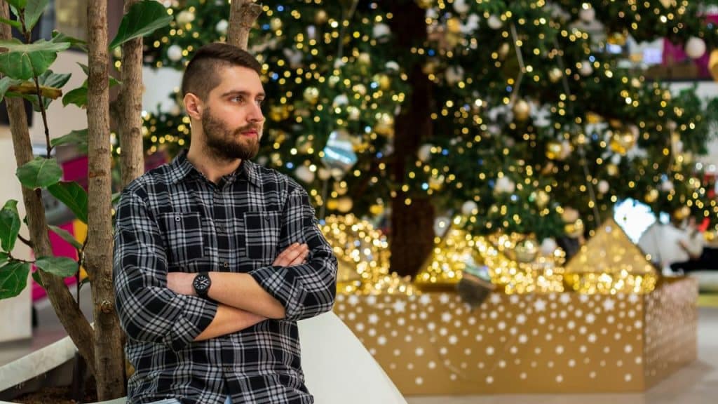 A man with crossed arms in a plaid shirt stands near a decorated Christmas tree.