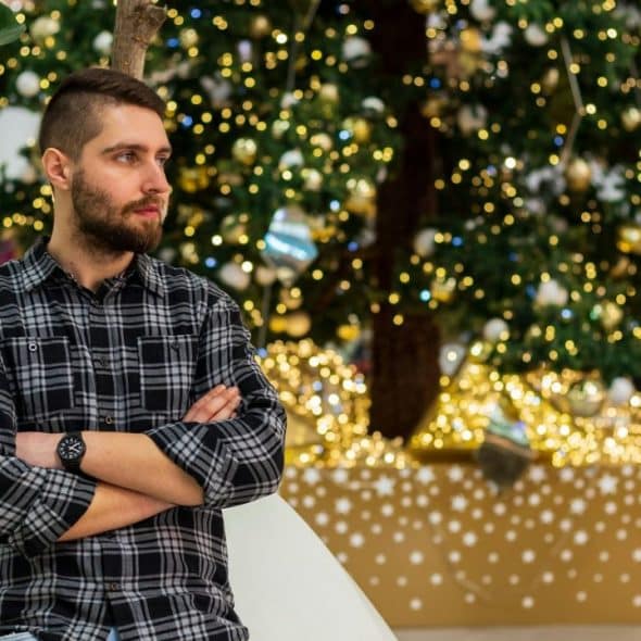 A man with crossed arms in a plaid shirt stands near a decorated Christmas tree.
