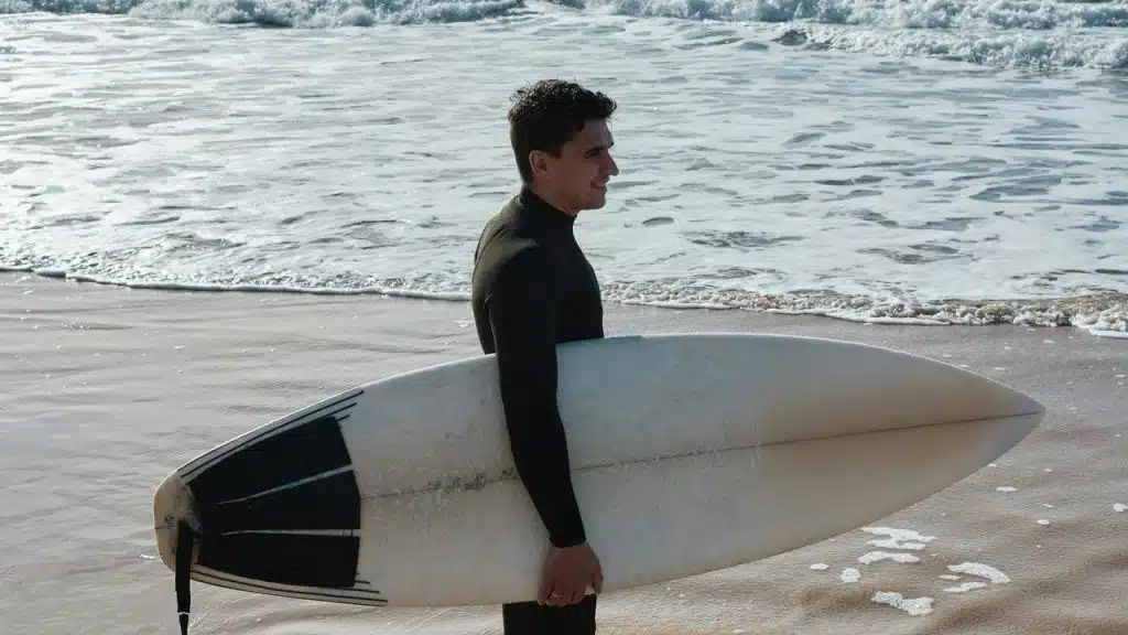 An adult in a wetsuit holding a surfboard by the shore.
