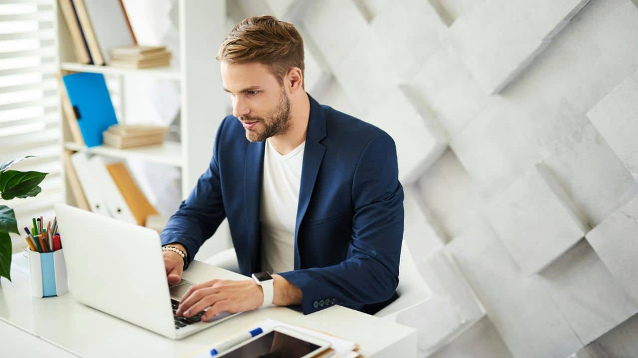 A man in a blue suit jacket working on a laptop at a white office desk.