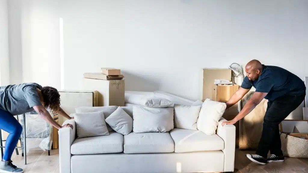 A couple moves a white couch in a room filled with moving boxes.