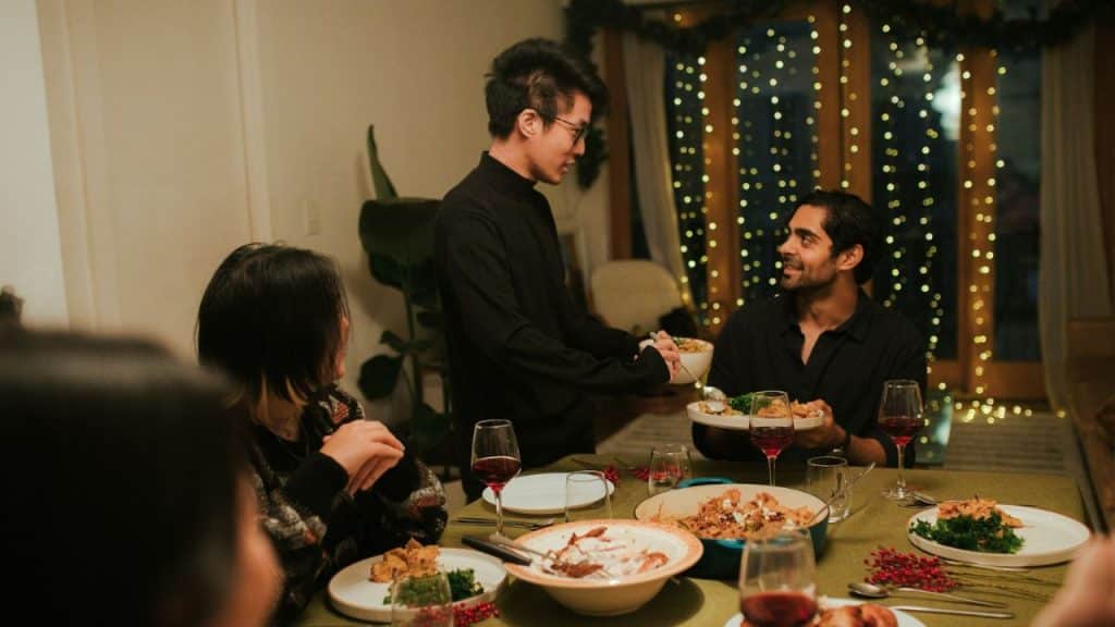 Man in black serving food to a smiling man seated at a dinner table with others.
