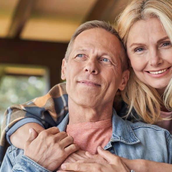 Middle-aged couple embracing and smiling outside, wearing casual denim and plaid shirts.