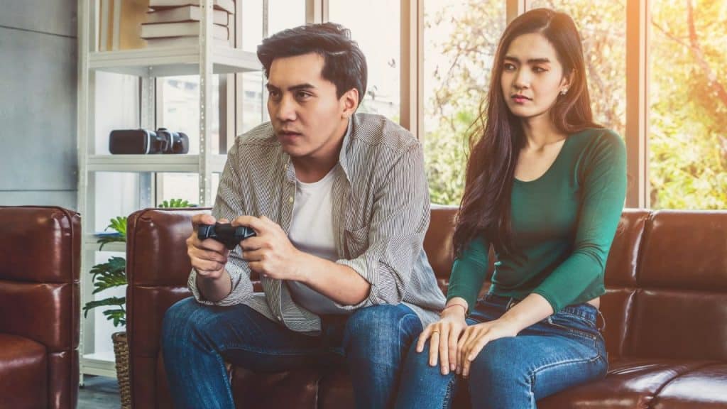 Man playing a video game on a couch while a woman sits next to him looking annoyed.