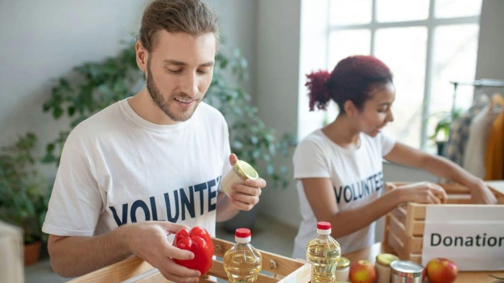 Man and woman in volunteer shirts sort donations, holding a pepper and canned goods.