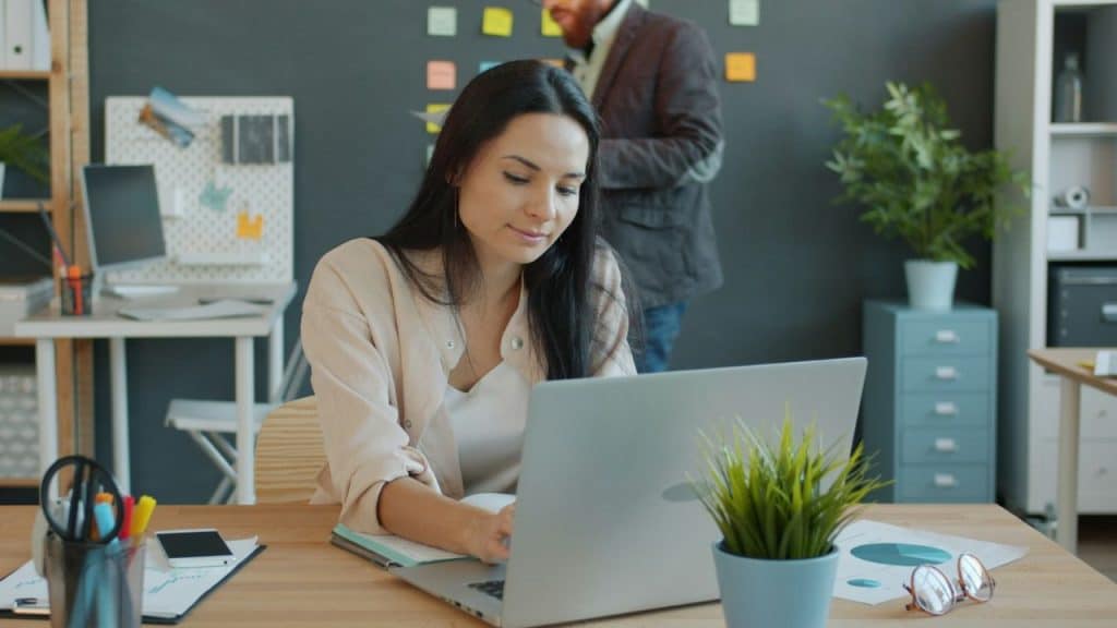 Woman working on a laptop at a desk in an office with a man standing behind her.