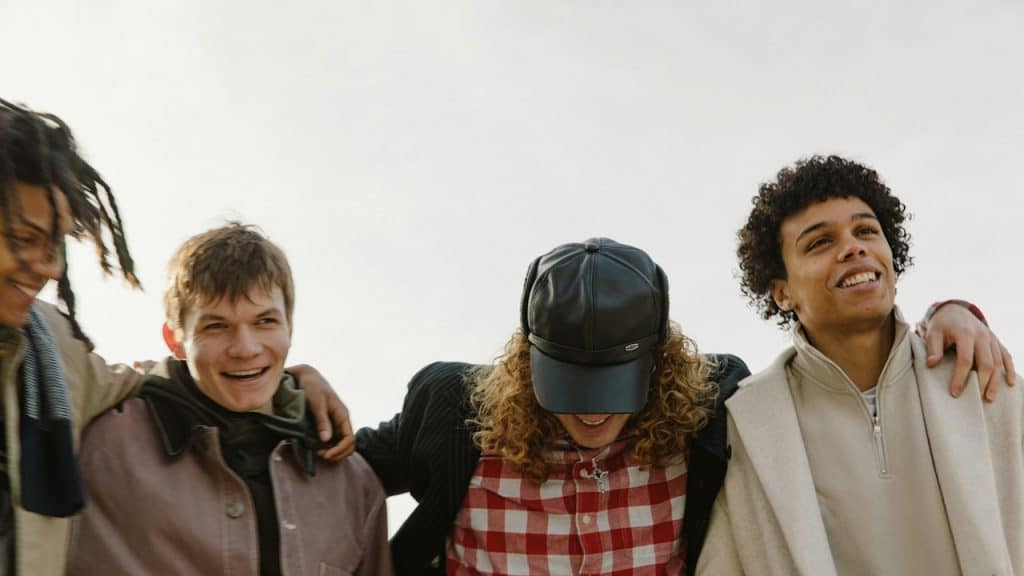A group of four young people standing side-by-side with their arms around each other's shoulders, smiling outdoors.