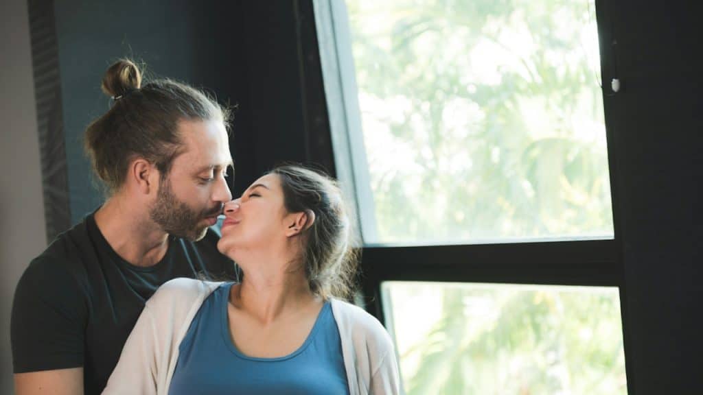 A couple is kissing by a window with a bright, lush green background outside.