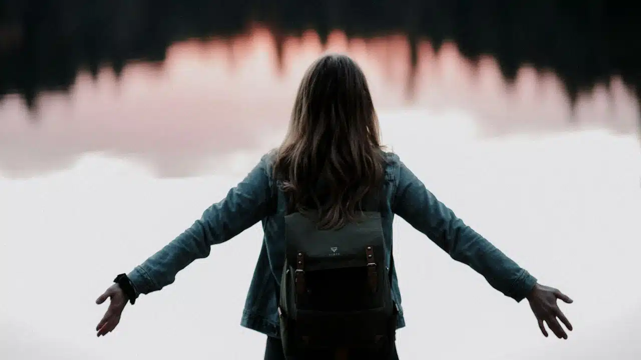 A woman standing is extending her arms while facing a body of water.