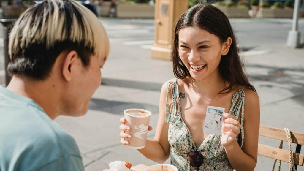 A woman laughing and showing a photo while sitting with a man at an outdoor café.