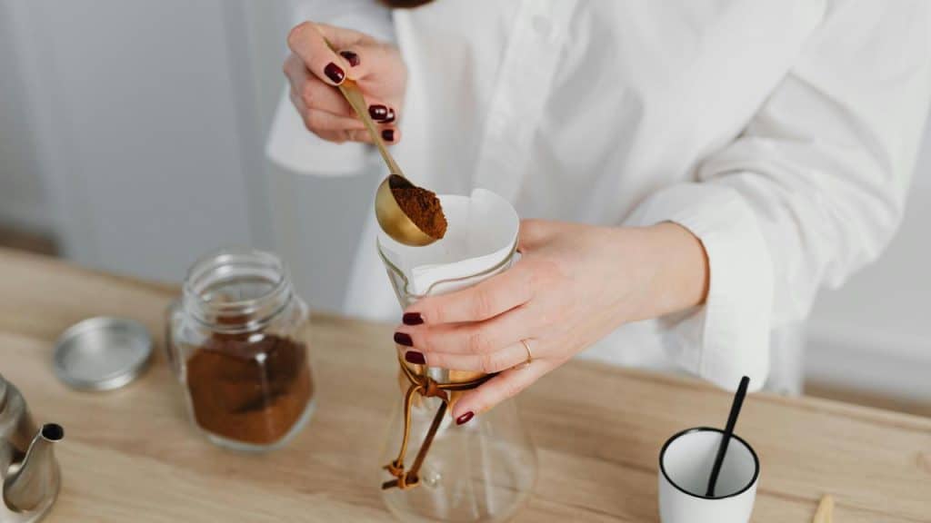 A person spooning ground coffee into a filter over a glass brewer.