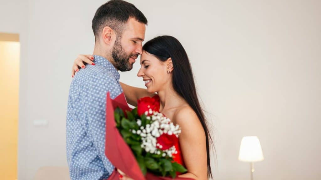 Man and woman embracing indoors; the woman is holding a bouquet of red and white flowers.