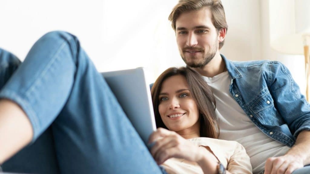 Woman lies across a man's lap; both look at a tablet together and smile.