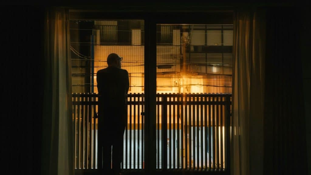 Silhouette of a person standing on a balcony at night, looking out at a brightly lit building across the way.