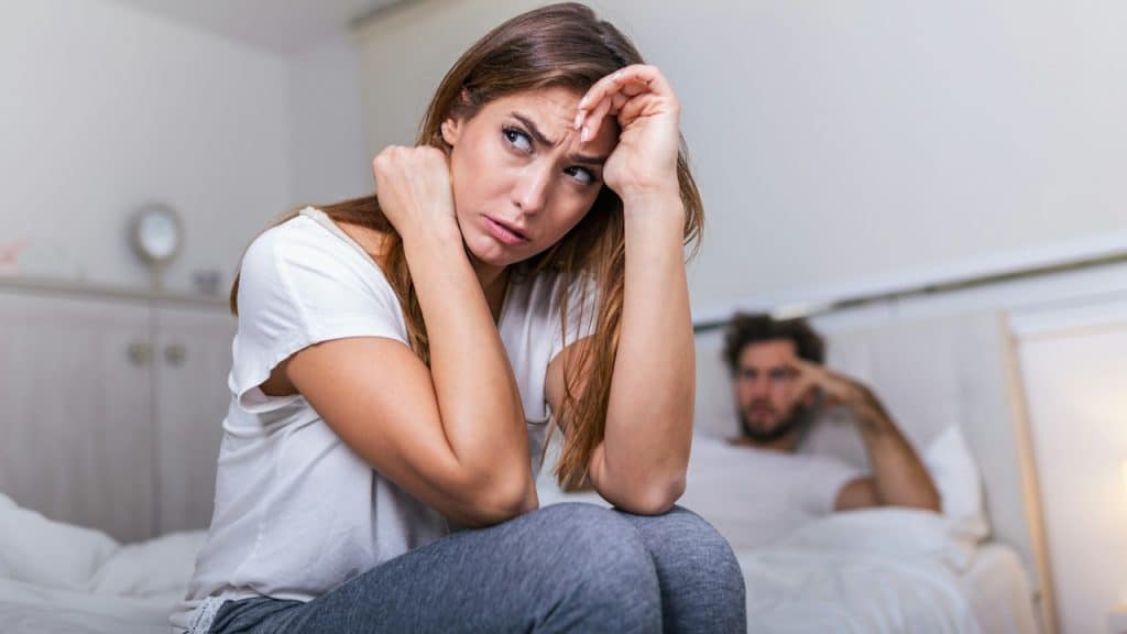 A woman sitting on the edge of the bed while her husband is leaning his back to the other side of the bed.