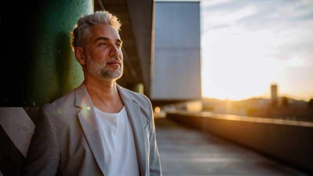 A gray-haired, bearded man stands outside at sunset, looking into the distance.