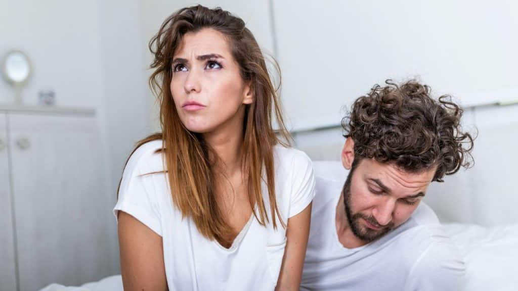 A woman sits on a bed with a troubled expression while a man looks down beside her.
